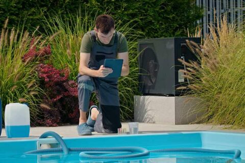 Pool technician checking data on a tablet beside a swimming pool with chemical containers and lush greenery.