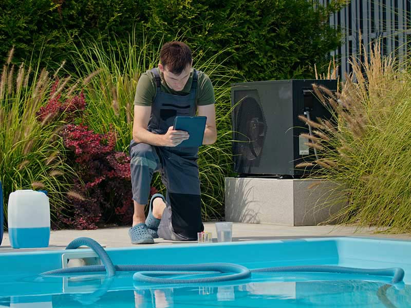 Pool technician checking data on a tablet beside a swimming pool with chemical containers and lush greenery.